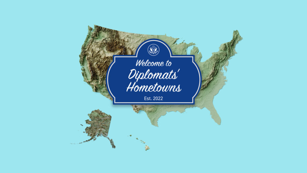 topographic map of United States over a light blue background, in foreground a dark blue sign with white text reads "Welcome to Diplomats' Hometowns Est. 2022"