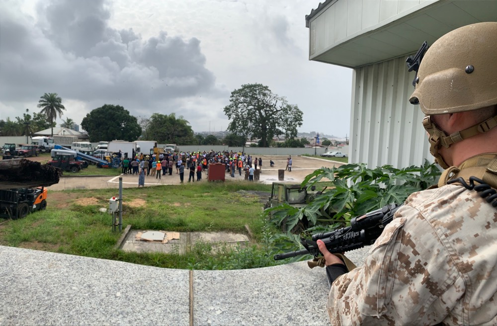 Regional Security Officer Michael Blees addresses the embassy community (gathered in the distance) during a fire drill at the U.S. Embassy as a Marine Security Guard provide security (right foreground), Monrovia, Liberia, March 2021. (U.S. Department of State photo)