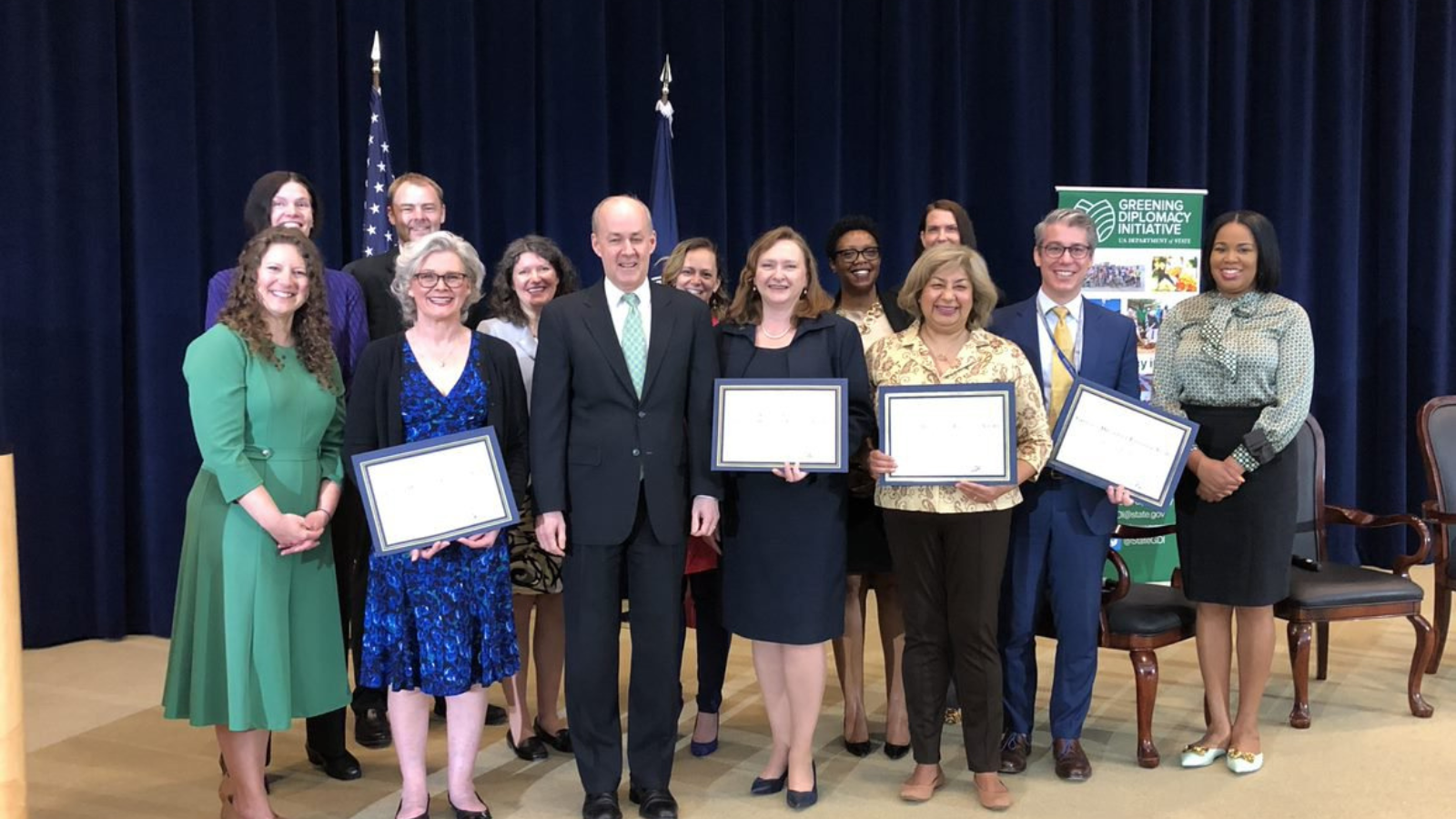 Deputy Secretary for Management and Resources McKeon stands with the winners of the 2022 Greening Diplomacy Initiative Awards, four individuals are holding certificates and an American and Department of State flag are featured in the background.