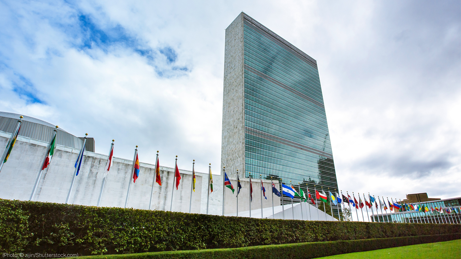 A tall, mirrored building reflects a backdrop of clouds, with a row of flag poles in front bearing the flags of the United Nations member countries.