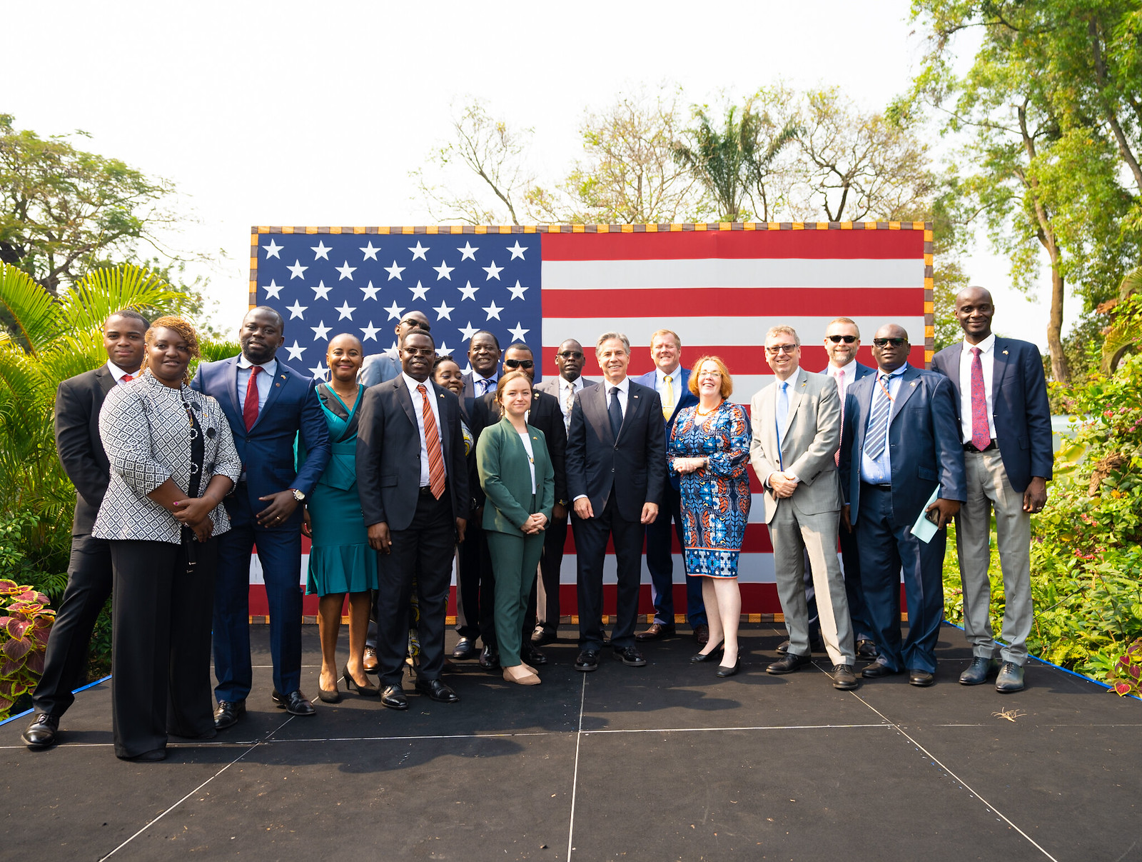 Secretary of State Antony J. Blinken poses for a photo with employees and families from U.S. Embassy Kinshasa in Kinshasa, Democratic Republic of the Congo. They are all standing in front of a large U.S. flag with trees on both sides.