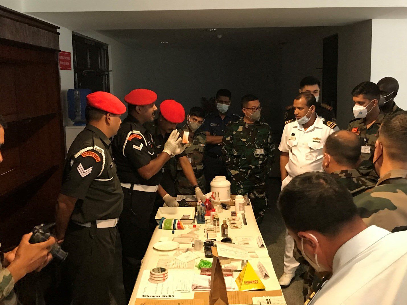 A group of Indian military police in red berets stand around a table and show their investigation kits to military and police trainers.