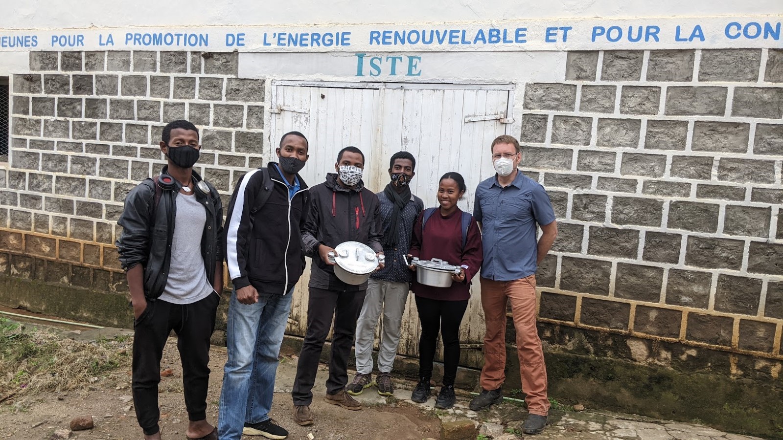 Six people stand in front of a wall. Two people hold pots and two people wear face masks.