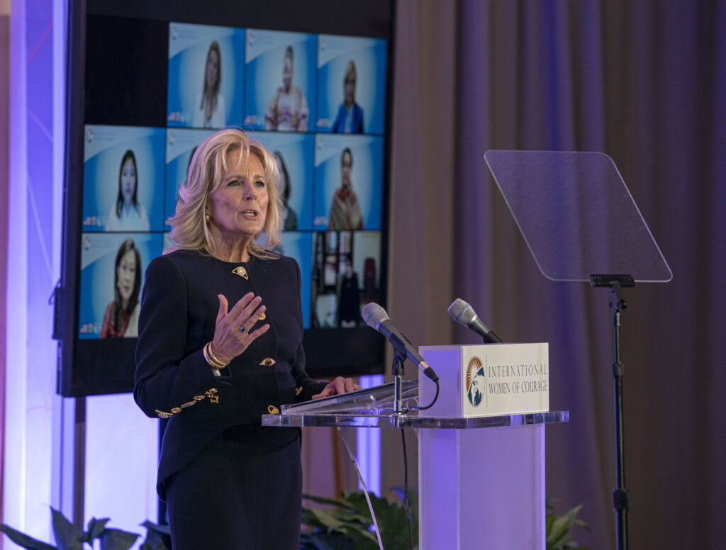 Dr. Biden speaks at International Women of Courage branded podium, video meeting of women on screen behind her