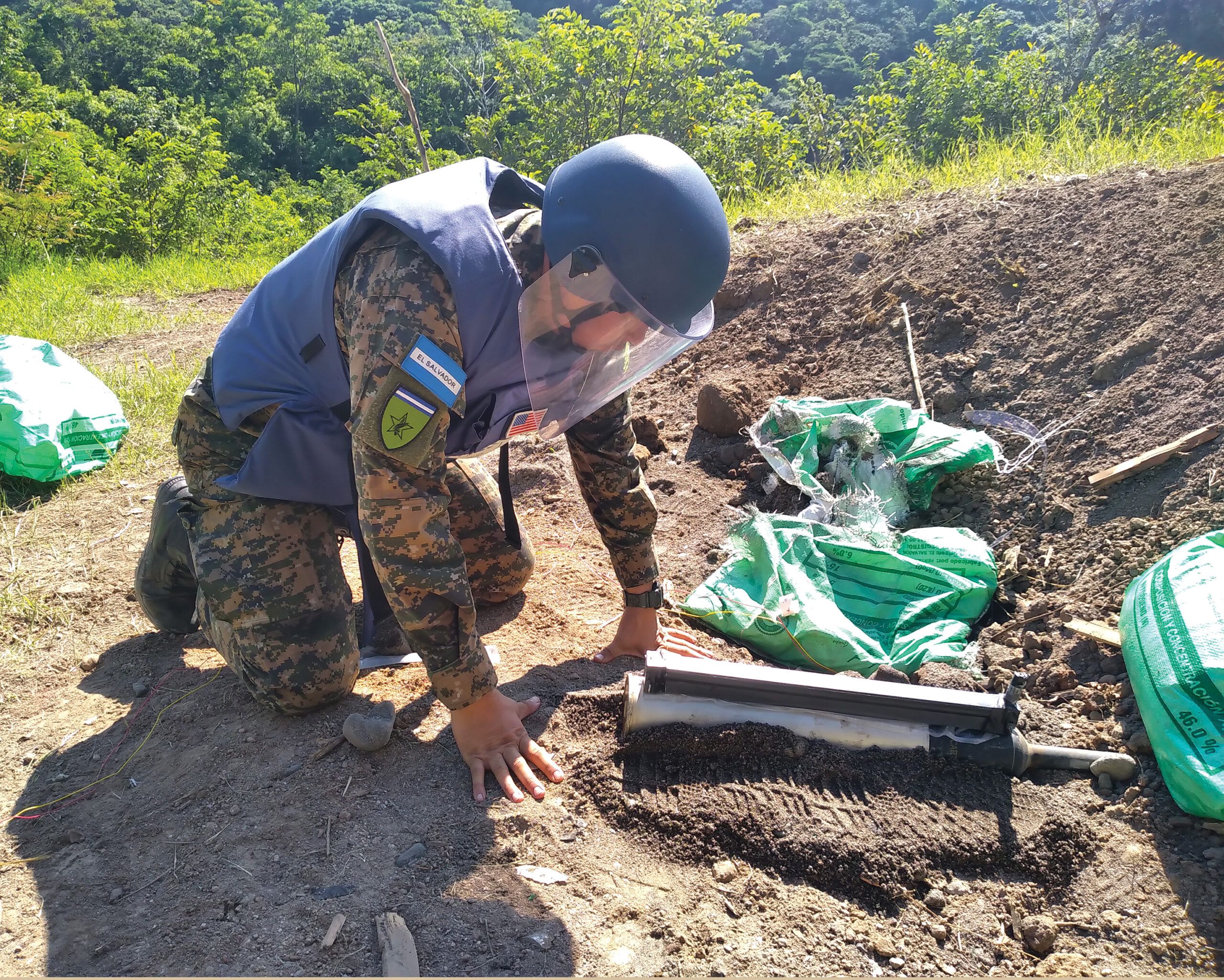 A man in protective gear kneeling in the dirt beside a metal object