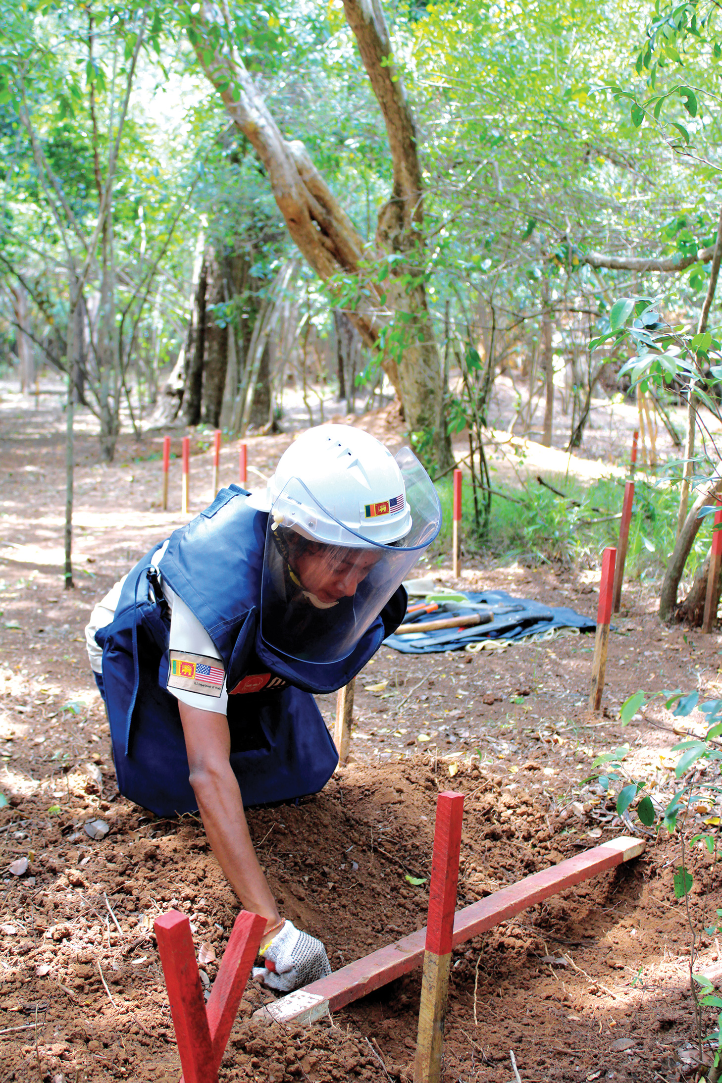 A kneeling person in protective gear with a tool in her hand digging in the dirt