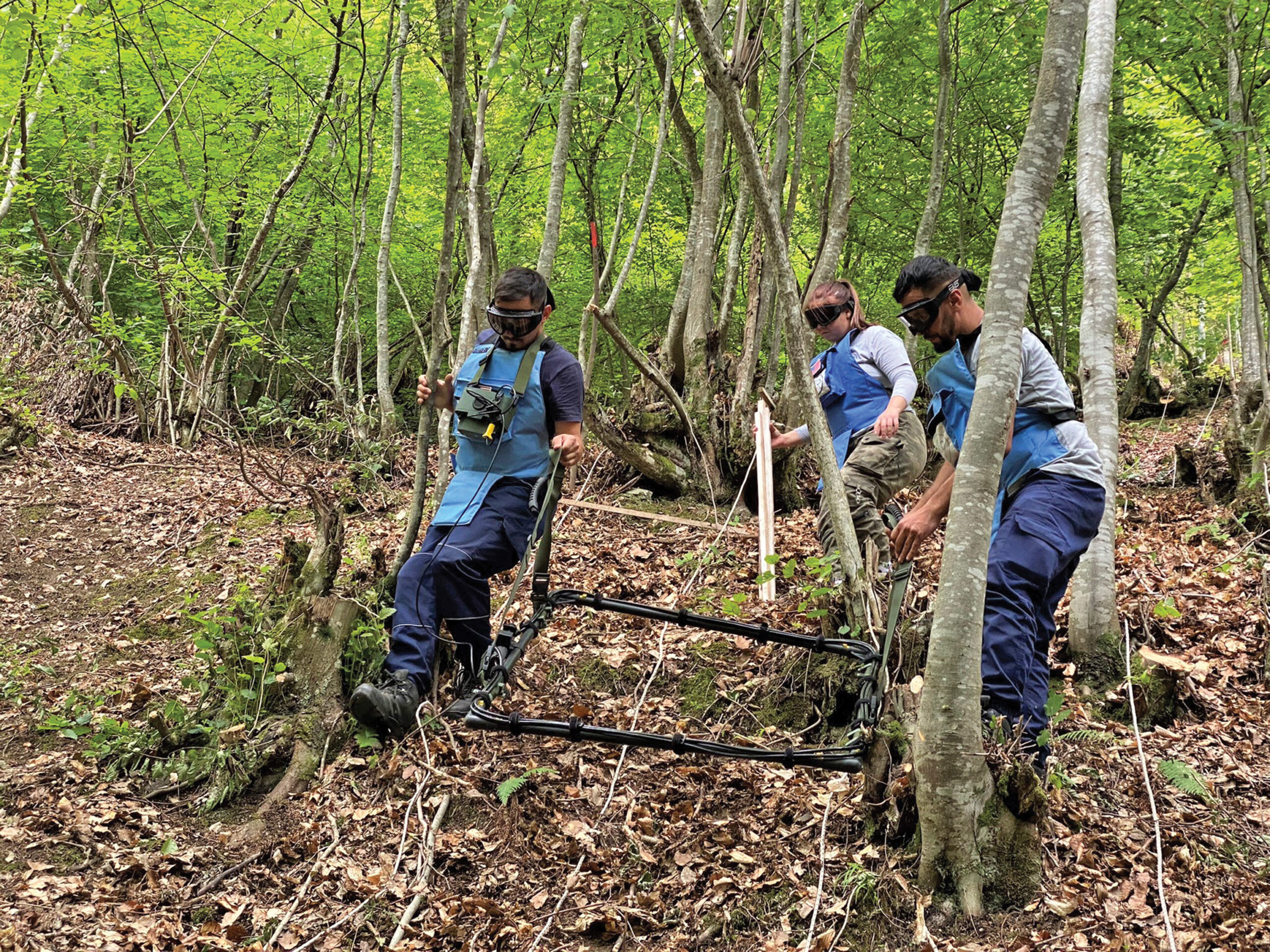 Three people in protective hear carry a large loop slightly above the ground through wooded land