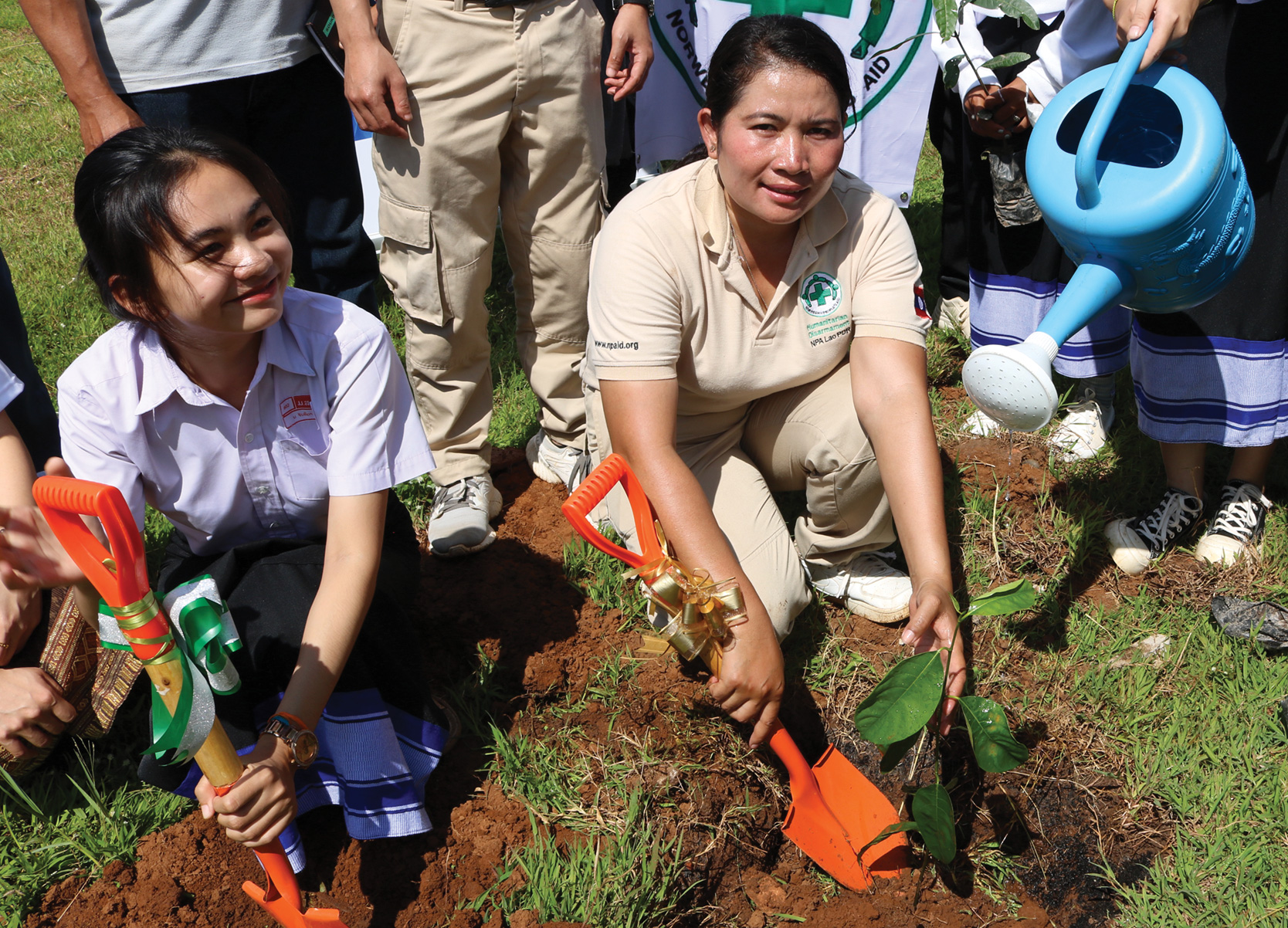 Two women with shovels squatting down to dig small holes in the ground to plant small trees.
