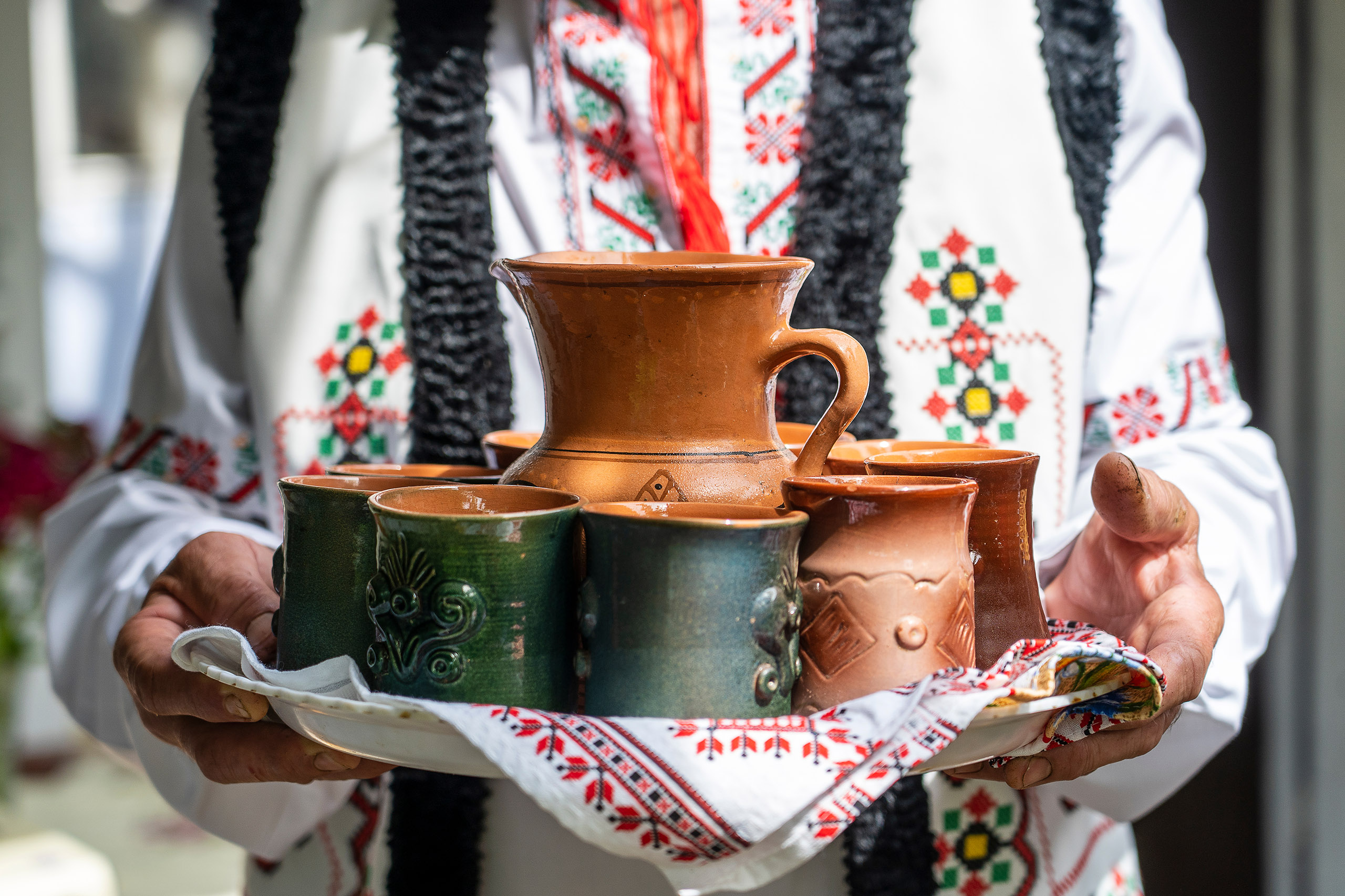 Moldovan man greets dear guests and invites them to drink homemade wine, close up. The hands of a man holding an earthenware jug with wine and glasses on a tray