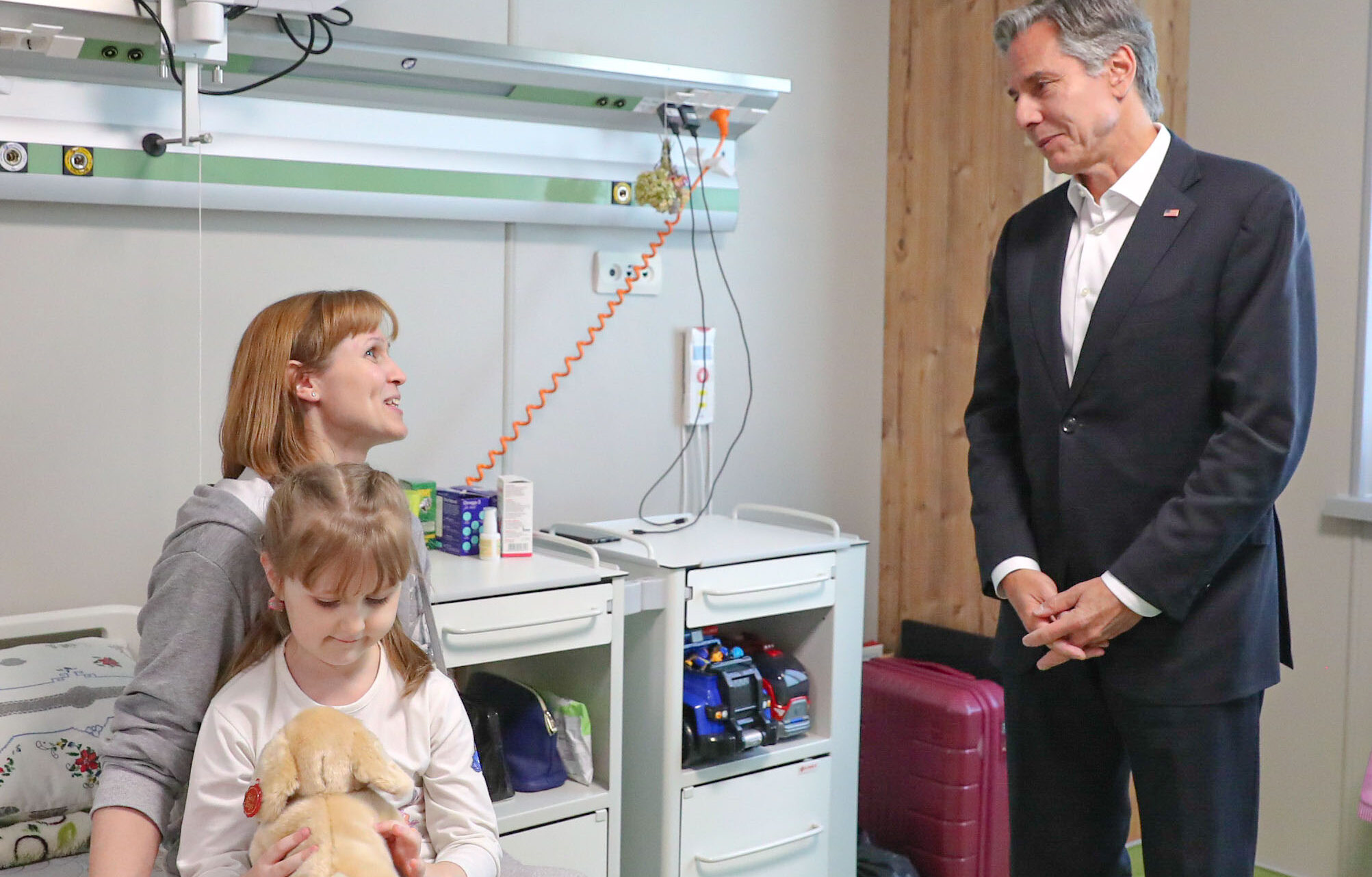 Secretary Blinken visits a family receiving care at a hospital in Ukraine in September 2022. He is standing and speaking with an adult and a child who are seated on a hospital bed.