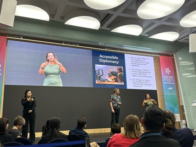 Angela Cannella is signing on stage with Robb Dooling and a Korean Sign Language interpreter, below the words “Accessible Diplomacy”