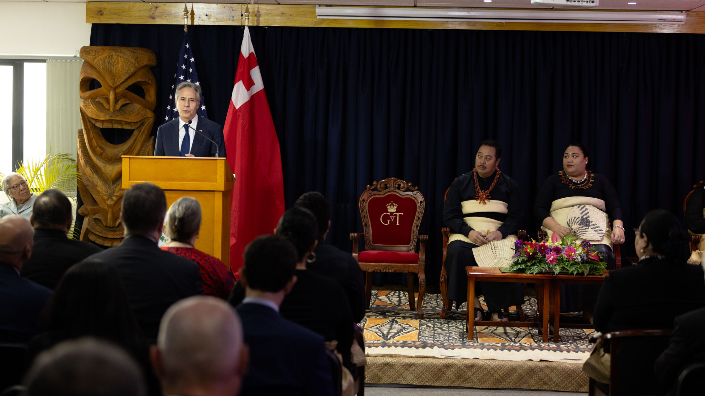 Secretary Blinken speaks from behind a lectern with the flags of the United States and Tonga behind him. Tonga's Crown Prince Tupouto'a 'Ulukalala and Crown Princess Sinaitakala are seated in chairs on stage. People in the audience are watching the dedication of the new U.S. embassy in Tonga, in Nuku'alofa