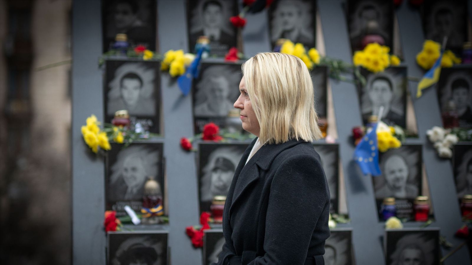 Ambassador Brink stands in front of a memorial wall with photos, candles, and flowers.