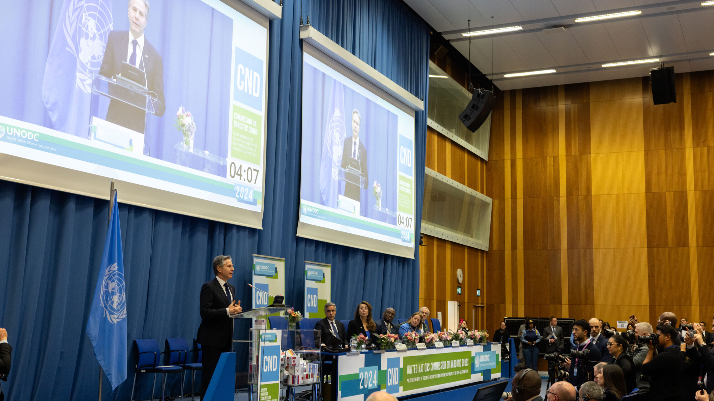 Secretary of State Blinken addressing the UN Commission on Narcotic Drugs in Vienna with his image appearing on two large screens behind him and an audience in front of him.
