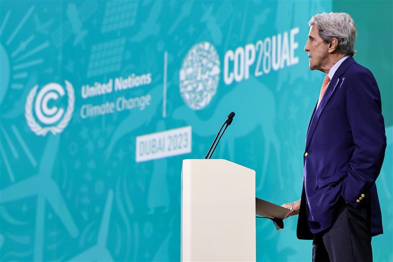 John Kerry standing at a podium next to banner that reads U.N. Climate Change COP 28 Dubai 2023. [State Department photo]